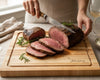 Person slicing a roasted beef roast on a Professional grade maple hardwood cutting board, with durable edge grain surface for carving meat, built for long-lasting use, includes personalization with The Johnsons branding. 