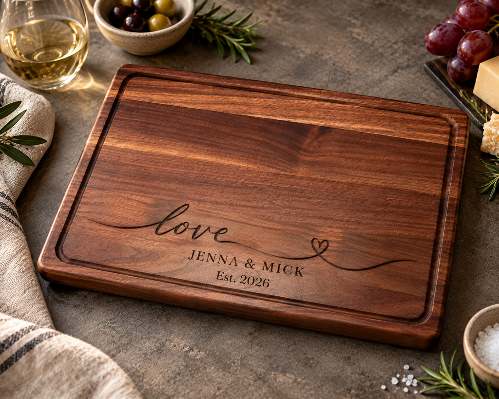 Personalized walnut wood cutting board with custom engraved names Jenna and Mick, Est. 2026, and elegant love design. Handcrafted hardwood chopping board with juice groove and non-slip rubber feet, displayed on a rich stone countertop. A thoughtful personalized wedding, anniversary, housewarming or bridal shower gift.