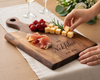 A personalized wooden charcuterie board engraved with 'THE Nekklas Est. 2025', styled on a tablecloth with cheese cubes, prosciutto, grapes, and rosemary, while a person's hands reach in to grab food.