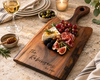 A personalized handled walnut wood charcuterie board engraved with The Robinsons Est. 2026, styled on a stone countertop with cheese, grapes, cured meat, figs, olives, honey, a glass of white wine, and a lit candle.