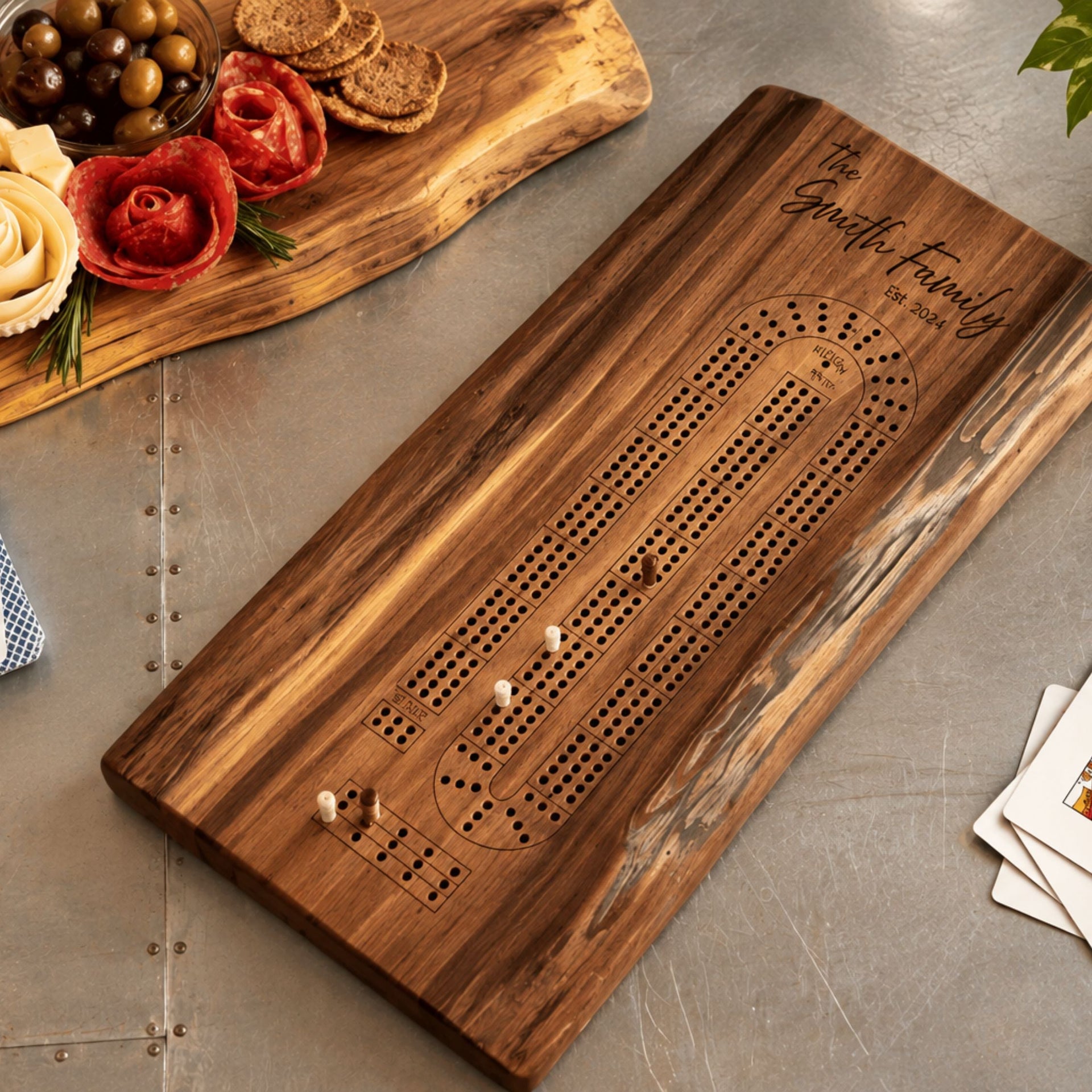 A personalized wooden live edge walnut cribbage board with engraved text reading 'The Smith Family' and a rustic live-edge serving board with decorative cheese and crackers, styled on a gray tabletop with playing cards nearby.