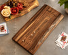 A personalized wooden live edge walnut cribbage board with engraved text reading 'The Smith Family' and a rustic live-edge serving board with decorative cheese and crackers, styled on a gray tabletop with playing cards nearby.