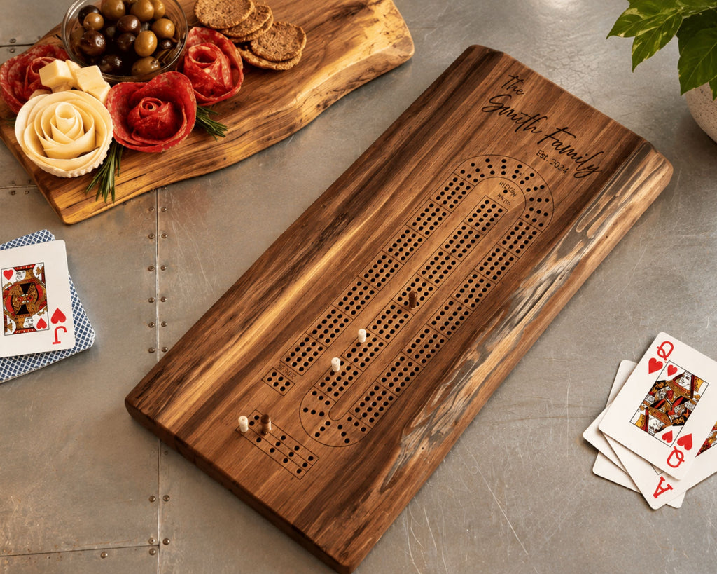 A personalized wooden live edge walnut cribbage board with engraved text reading 'The Smith Family' and a rustic live-edge serving board with decorative cheese and crackers, styled on a gray tabletop with playing cards nearby.