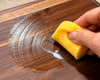 A close-up of a hand holding a yellow sponge and wiping a wooden surface with cutting board conditioner to enhance natural wood grain, restore rich colour, and revive dull wood