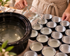 A person in a beige apron is making board conditioner as she pours melted Beeswax and mineral oil from a glass measuring pitcher into multiple small round metal tins arranged on a table next to a large black pot.