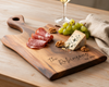 A personalized wooden charcuterie board engraved with 'The Robinsons EST. 2025' styled on a wooden table with sliced salami, a wedge of blue cheese, green grapes, walnuts, and a glass of white wine in the background.