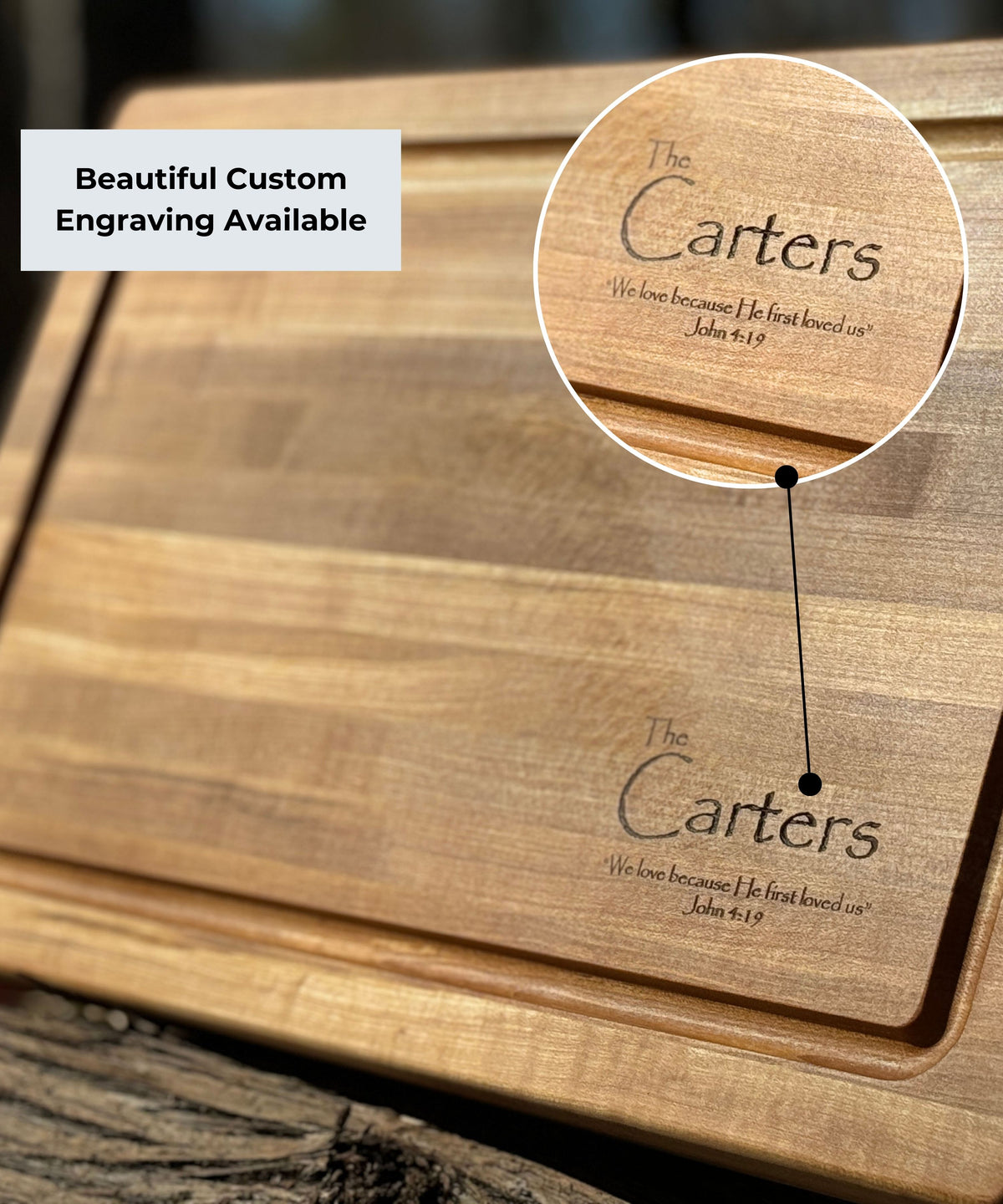 A beautifully crafted wooden cutting board with custom engraving that reads "The Carters" and includes the Bible verse "We love because He first loved us - John 4:19." The board's natural grain shines in a rustic outdoor setting.