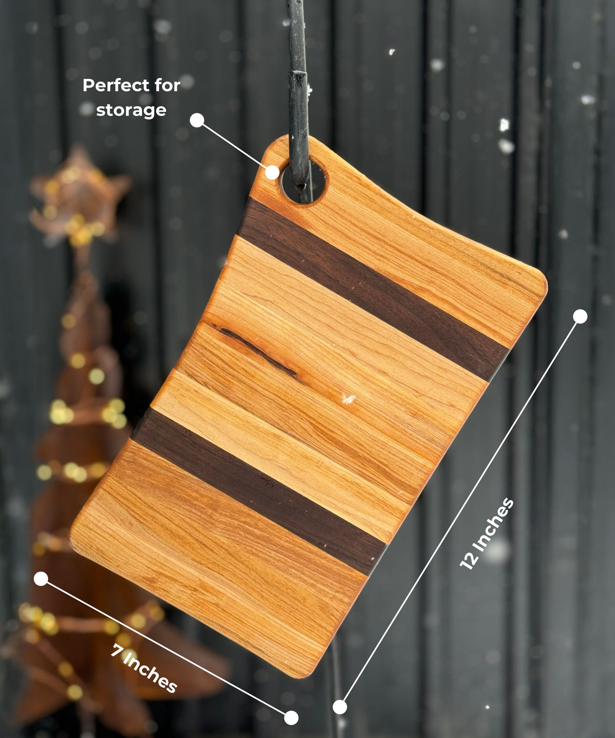 A 12-inch by 7-inch wooden cutting board with alternating maple and walnut wood stripes, hanging from a hook through a built-in hole for easy storage. The background features a dark wooden wall with snow falling and a blurred decorative wooden tree.