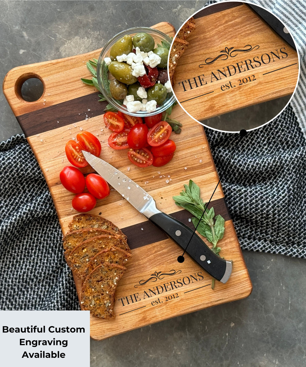 A personalized wooden cutting board featuring maple &amp; walnut stripes, engraved with The Andersons est. 2012. Topped fresh cherry tomatoes, olives with feta, herbs, &amp; crispy crackers. Text overlay reads Beautiful Custom Engraving Available.