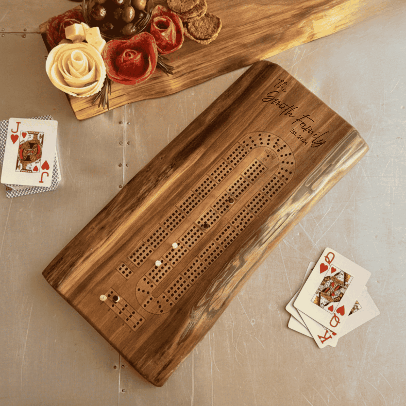 A live edge walnut solid wood cribbage board engraved with The Smith Family, Est. 2024 displayed alongside a live edge walnut charcuterie spread with cheese, meats, and olives. Cards and a wine glass complete the cozy, personalized game night setup