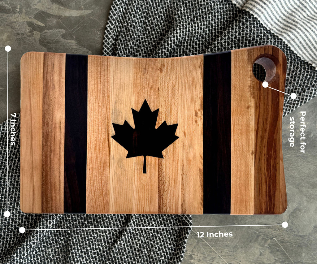 The striped maple and walnut canadian flag cutting board with a laser-engraved, epoxy filled maple leaf in the center. The board dimensions are illustrated 12 by 7 inches. a perfectly placed hole at the top meant for hanging.