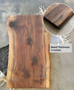 A live-edge walnut face grain slab charcuterie board with a rich wood grain, personalized engraving reading, Mr. & Mrs. Johnson 12.22.2021, and a close-up inset highlighting its 1.1-inch thickness. The board rests on a striped fabric backdrop.