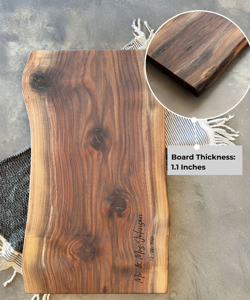 A live-edge walnut face grain slab charcuterie board with a rich wood grain, personalized engraving reading, Mr. & Mrs. Johnson 12.22.2021, and a close-up inset highlighting its 1.1-inch thickness. The board rests on a striped fabric backdrop.