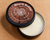 An open round metal tin of beeswax and mineral oil wood balm, showing the creamy off-white balm inside, with the lid leaning against it on a tan textured background. The lid features a wood-grain pattern with white text reading 'Board Butter' 100% food grade