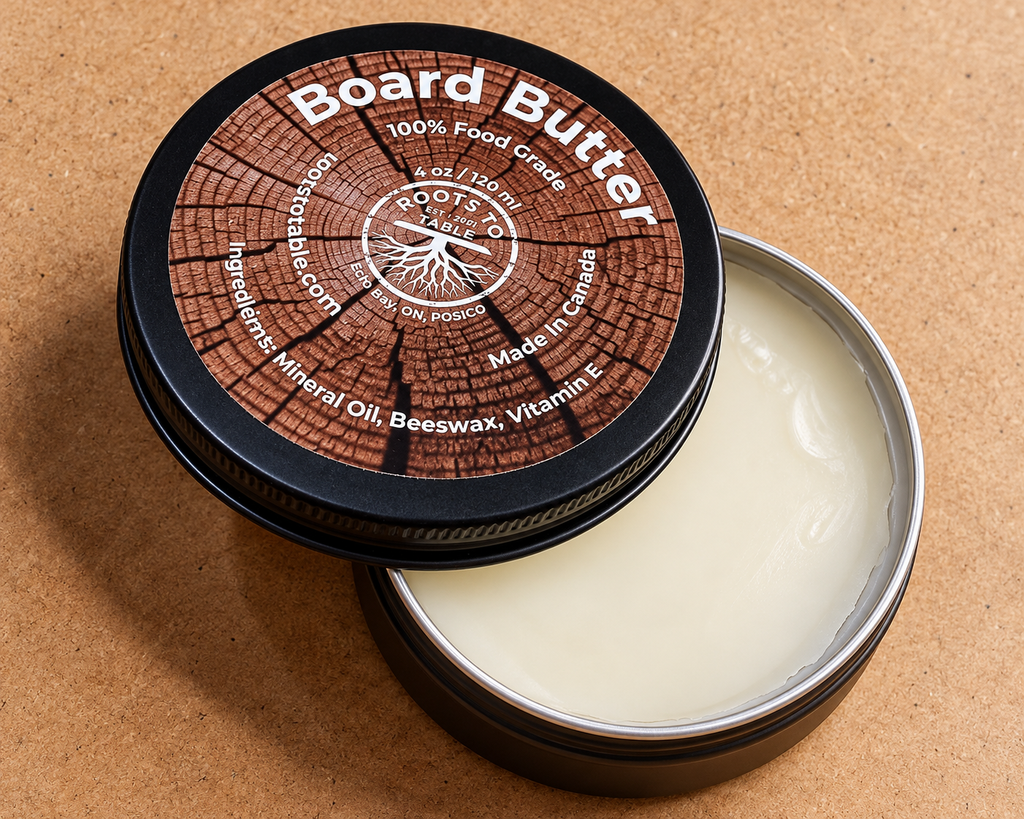 An open round metal tin of beeswax and mineral oil wood balm, showing the creamy off-white balm inside, with the lid leaning against it on a tan textured background. The lid features a wood-grain pattern with white text reading 'Board Butter' 100% food grade