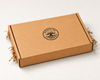 A square brown kraft cardboard box with a black printed circular logo reading 'ROOTS TO TABLE' in the center, photographed on a clean white background with crinkle paper filler visible at the sides. This is a gift ready personalized charcuterie board