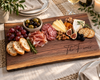 A personalized wooden charcuterie board engraved with 'The Ferris' and 'Marie & Sean Nicole 2025' is styled on a woven placemat with bread, grapes, prosciutto, cheese, nuts, figs, olives, and crackers. A wine glass and candle are softly blurred in the background.