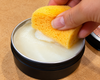 A close-up of a hand dipping a yellow sponge into a black tin of Food-safe cutting board conditioner used to Nourish dry wood, condition wood fibres, replenish moisture-resistant finish.