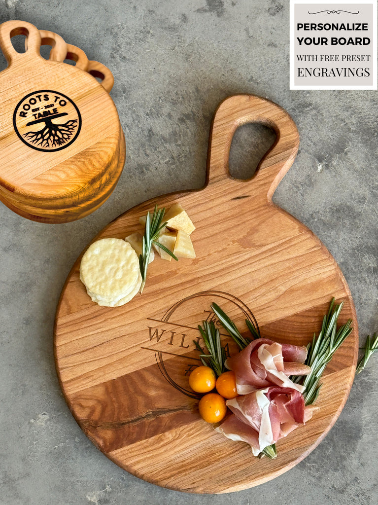 Round wooden charcuterie board with a handle, engraved. Styled with crackers, cheese, prosciutto, yellow tomatoes, and rosemary sprigs. Accompanied by 4 stacked wooden coasters featuring the Roots to Table logo, set against a textured stone backdrop.