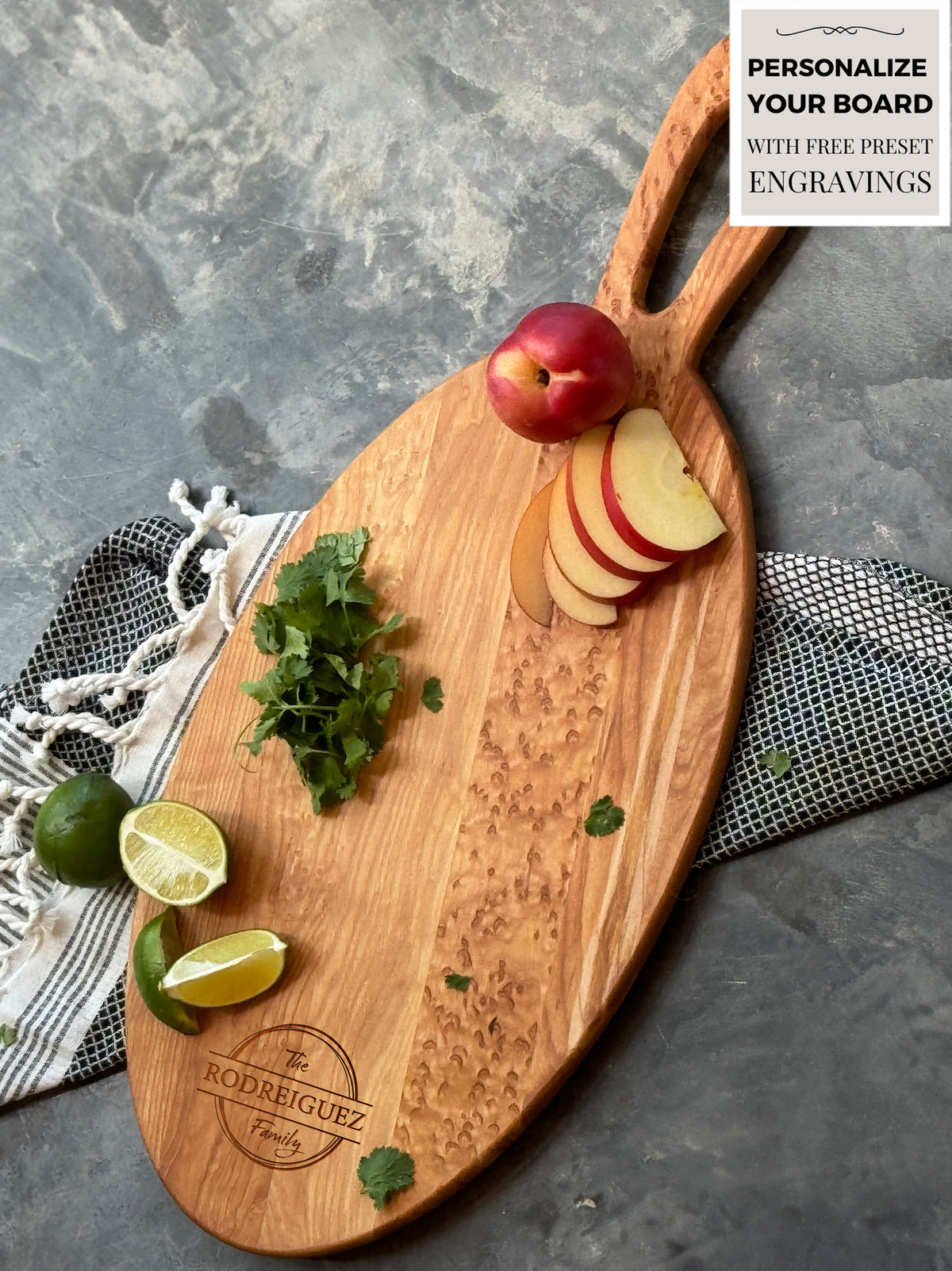 24 Inch Oval maple wood charcuterie board with a handle, featuring engraved customization for The Rodriguez Family. Displayed with sliced apples, lime wedges, and fresh cilantro on a textured surface, accented by a striped fabric towel.
