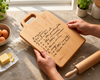 A light maple wood cutting board with a handle and handwritten recipe engraved in french, held by two hands over a floured kitchen counter with a rolling pin, eggs, butter, and small potted plants in the background. Makes a perfect gift.