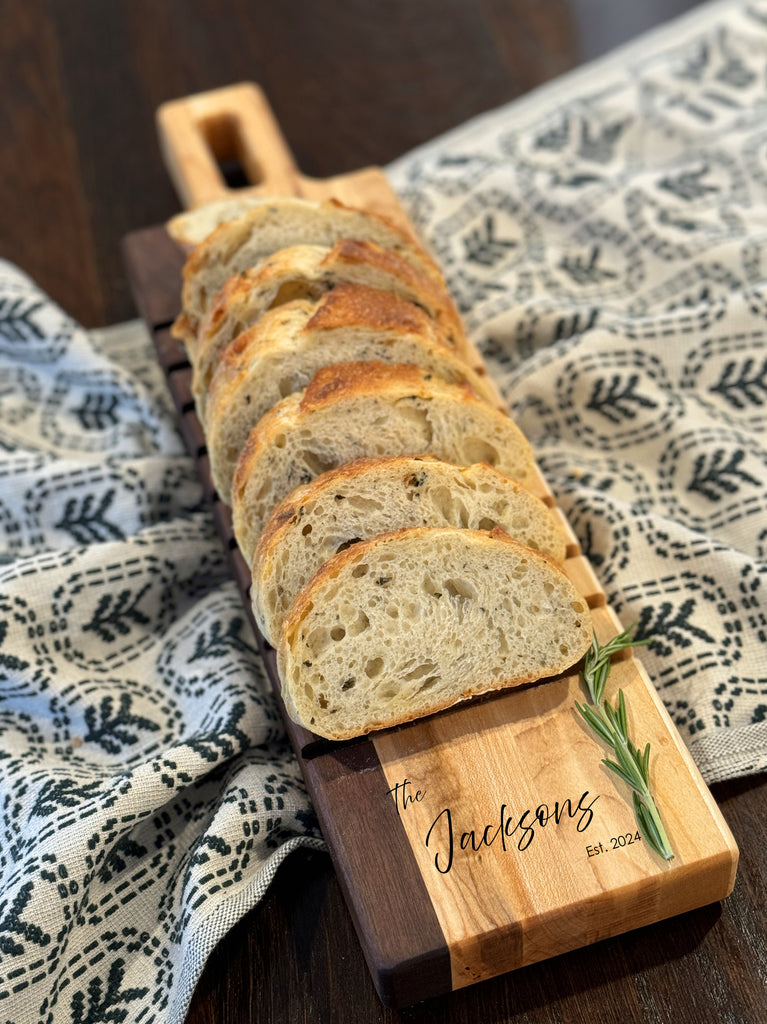 A beautifully crafted walnut breadboard featuring The Jacksons engraving with established date. Slices of fresh bread rest on the board, paired with sprigs of rosemary, set against a patterned cloth backdrop. Perfect for elegant serving.