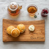 Wooden cutting board with scones, clotted cream, and jam, accompanied by a teapot and cup of tea on a light surface.