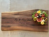 A live-edge walnut charcuterie board featuring a natural grain pattern, personalized with Robertson Family engraving. Topped with crackers, olives, salami roses, and cheese, it rests on a silver metal coffee table from restoration hardware.