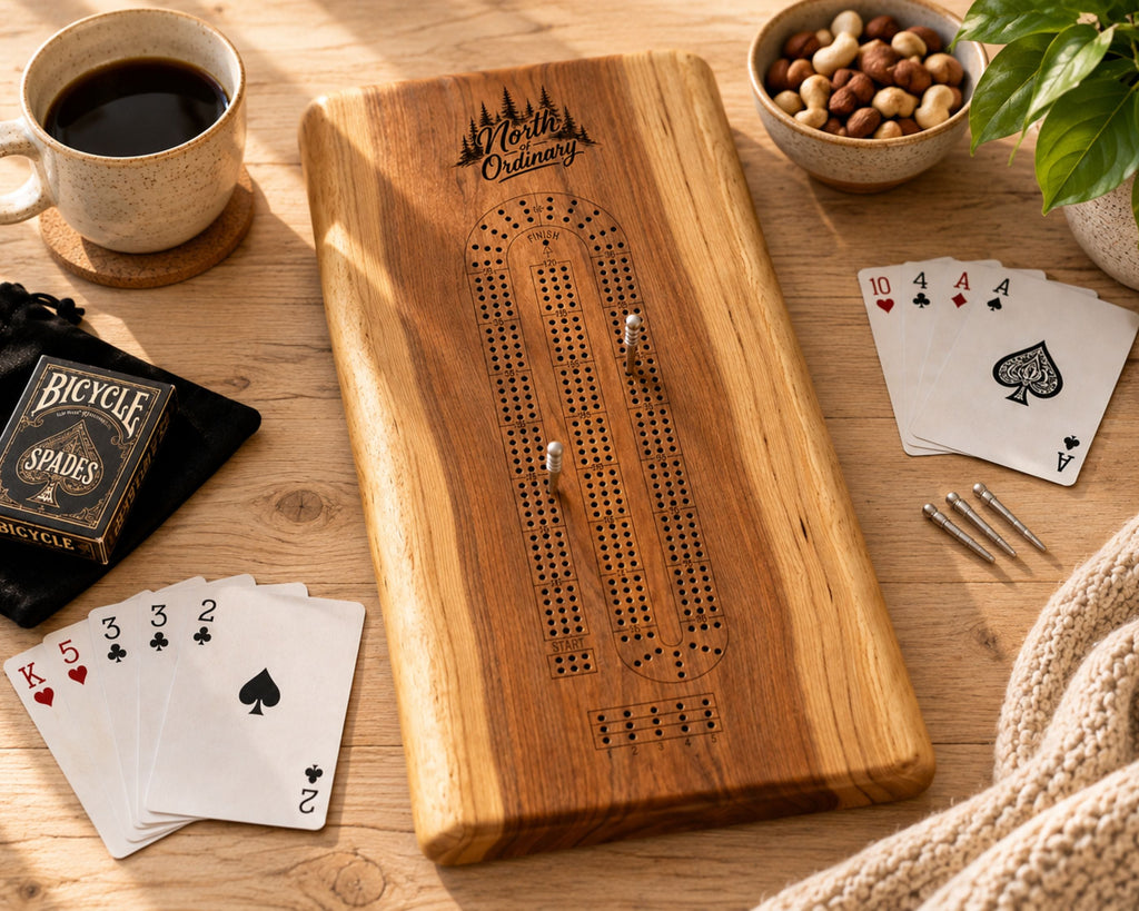 A personalized wooden live edge cribbage board set on a wooden table with playing cards, a cup of coffee, nuts, and a knit blanket. The board has an engraved logo for 'Keller's Outfitters' and a Bicycle Spades deck box is visible on the left.