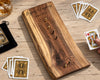 A wooden peg-and-hole cribbage board set on a light wood tabletop, with personalized engraving text reading 'Pegging & Laughing since 1972.' Playing cards are scattered around it, including a visible deck bag and a hand holding a card.