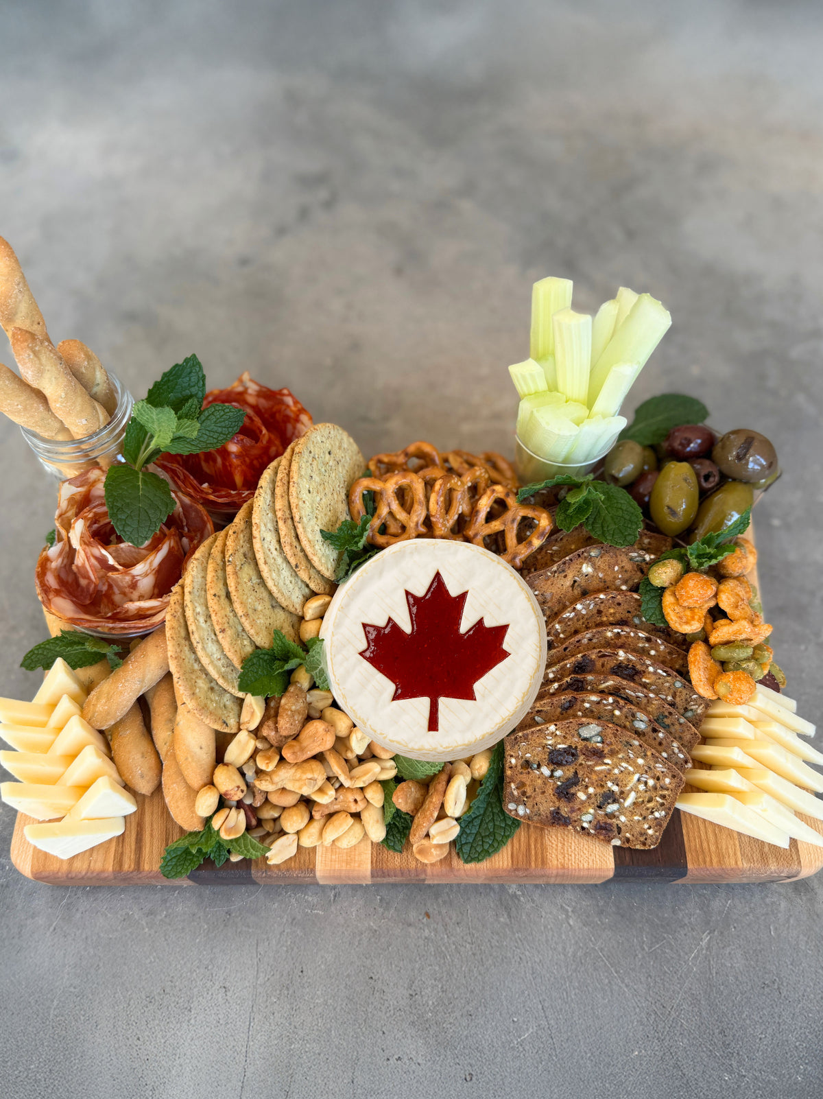 The canadian flag cutting board being used as a charcuterie board featuring assorted crackers, cheeses, meats, nuts, olives, celery sticks, and a wheel of brie topped with a red maple leaf design made of jam.