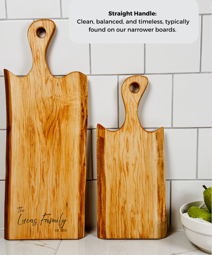 Two handcrafted Live Edge maple serving boards displayed upright, featuring natural wood grain and engraving. The charcuterie boards are more narrow that the wider options and have a straight handle.
