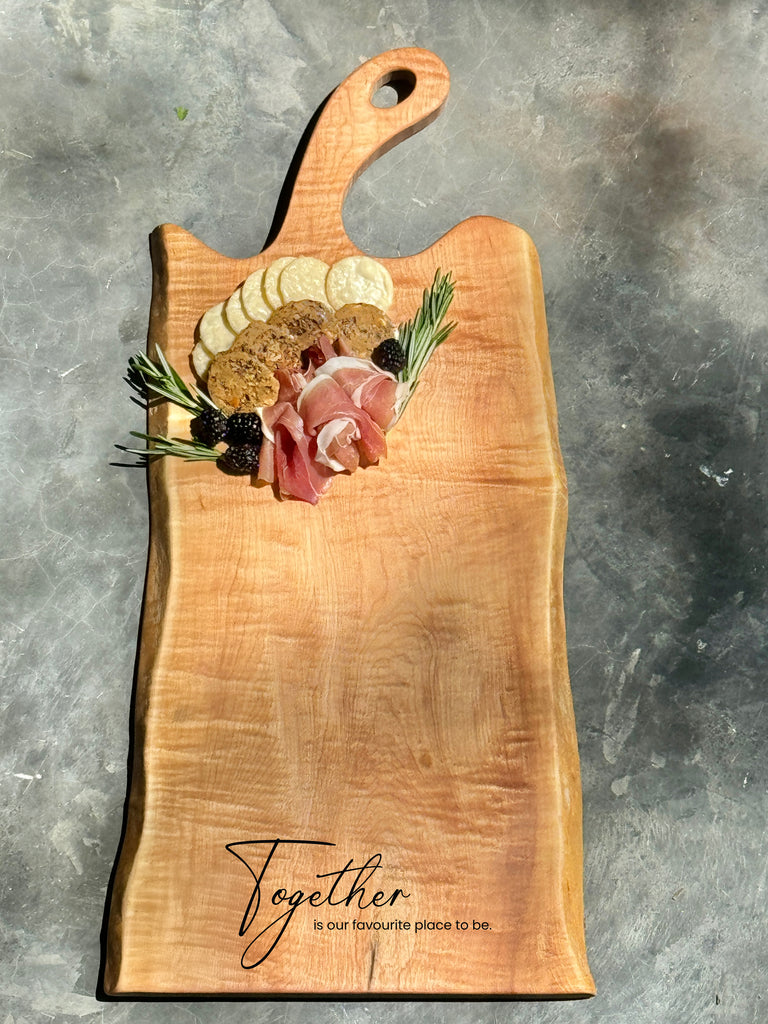 24-inch maple live edge charcuterie board with a curved handle, beautifully arranged with cheeses, meats, and fruit, featuring an engraving that reads “Together is my favourite place to be.”