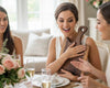Three women seated around a dining table in a bright, elegant room at a bridal shower. The central woman holds a large wooden cutting board with handle that she has received as a wedding gift.