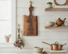 A rustic wooden cutting board with a handle, featuring the engraved text 'The Kippsing House', hanging on a white shiplap wall in a styled kitchen setting.