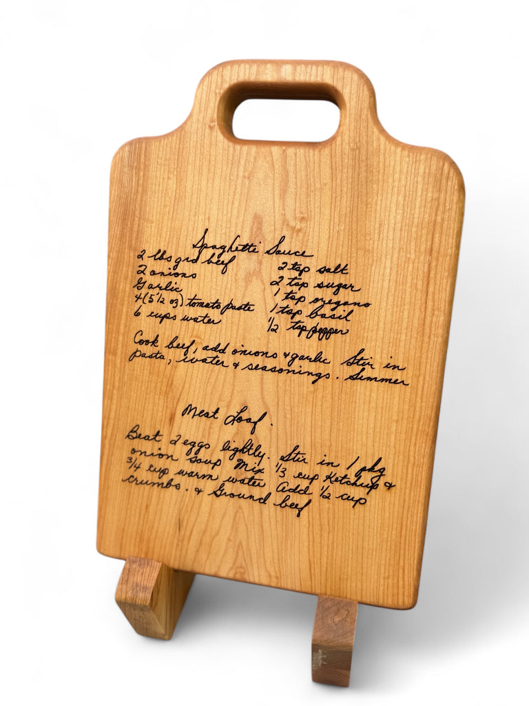 two hand written recipes engraved on a face grain maple cutting board, the board sits on a two piece stand.