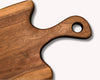A close-up of a wooden serving board or cutting board with a rounded handle and a circular hole, showing a warm medium-brown wood grain on a light gray background.
