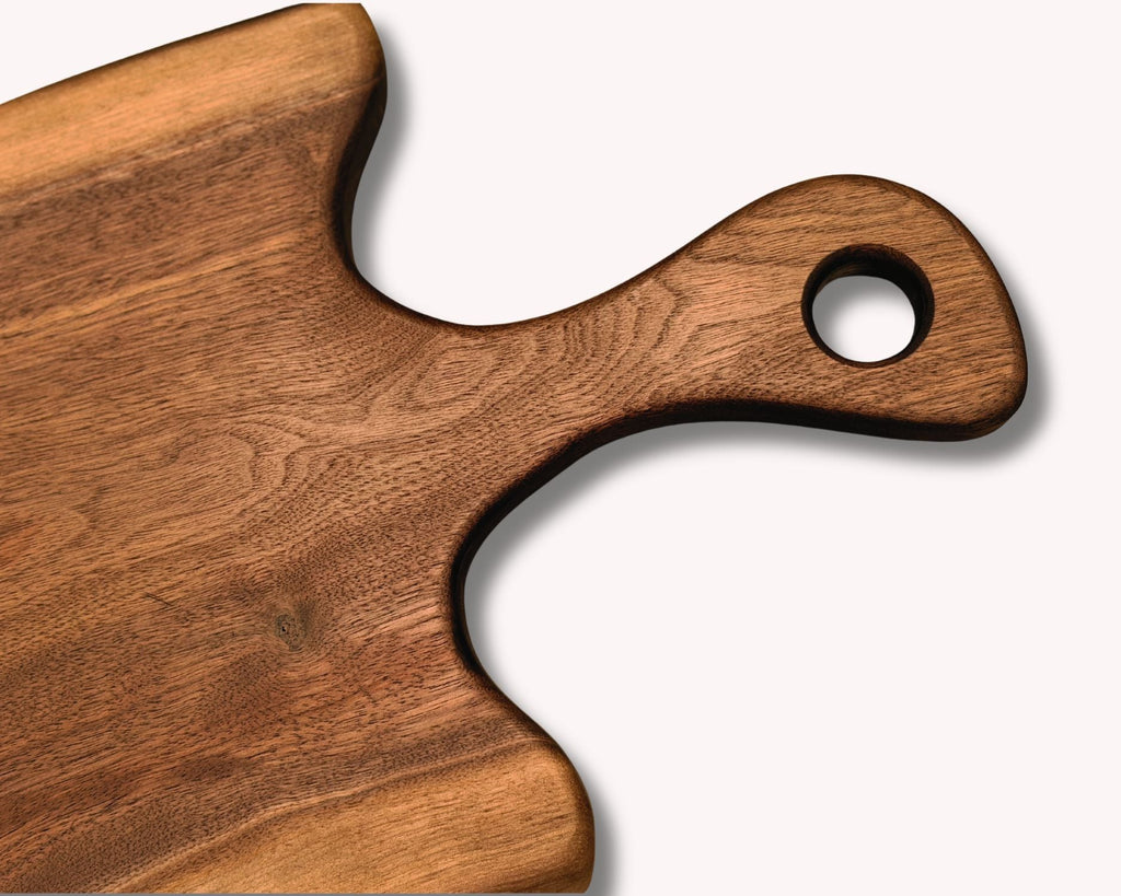 A close-up of a wooden serving board or cutting board with a rounded handle and a circular hole, showing a warm medium-brown wood grain on a light gray background.