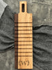A handcrafted breadboard made with maple and walnut, featuring horizontal grooves and a personalized engraving of the letter W, framed by laurel leaves. The board is displayed on a textured gray surface with striped and checkered cloth accents.