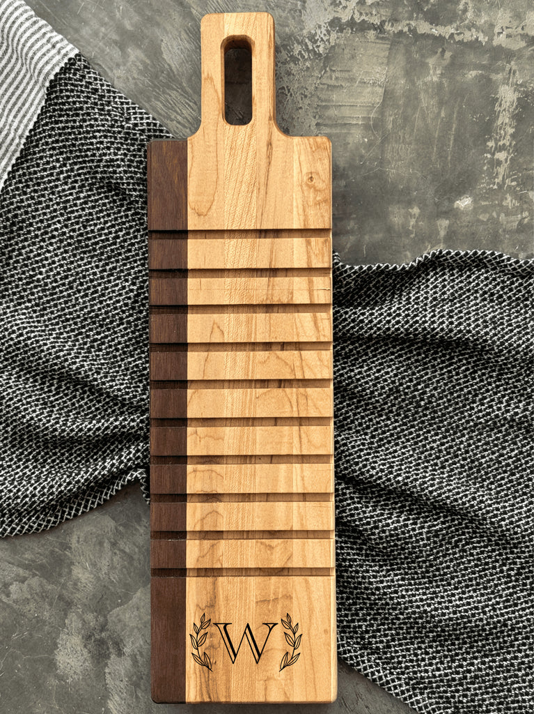 A handcrafted breadboard made with maple and walnut, featuring horizontal grooves and a personalized engraving of the letter W, framed by laurel leaves. The board is displayed on a textured gray surface with striped and checkered cloth accents.