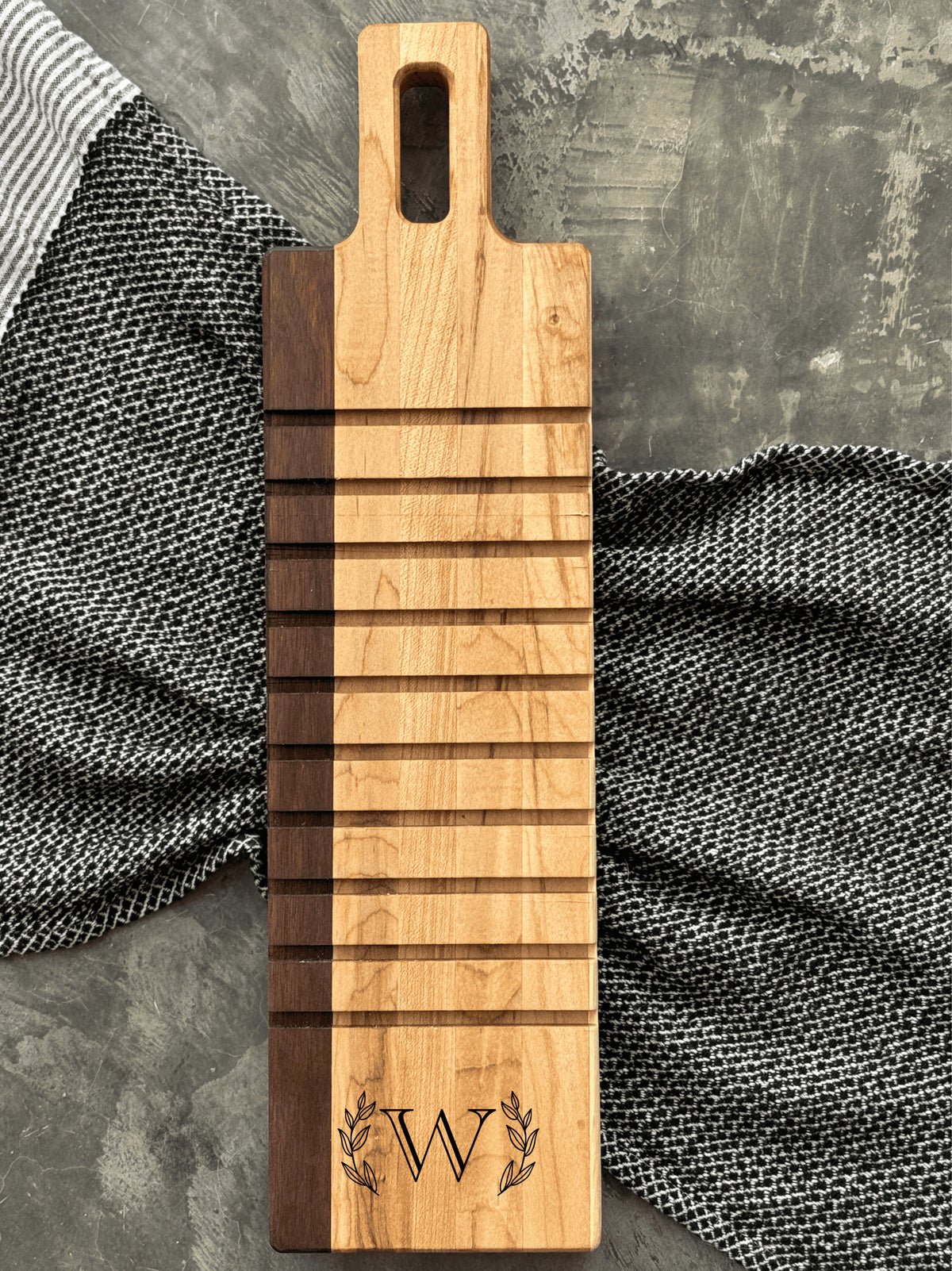 A handcrafted breadboard made with maple and walnut, featuring horizontal grooves and a personalized engraving of the letter W, framed by laurel leaves. The board is displayed on a textured gray surface with striped and checkered cloth accents.