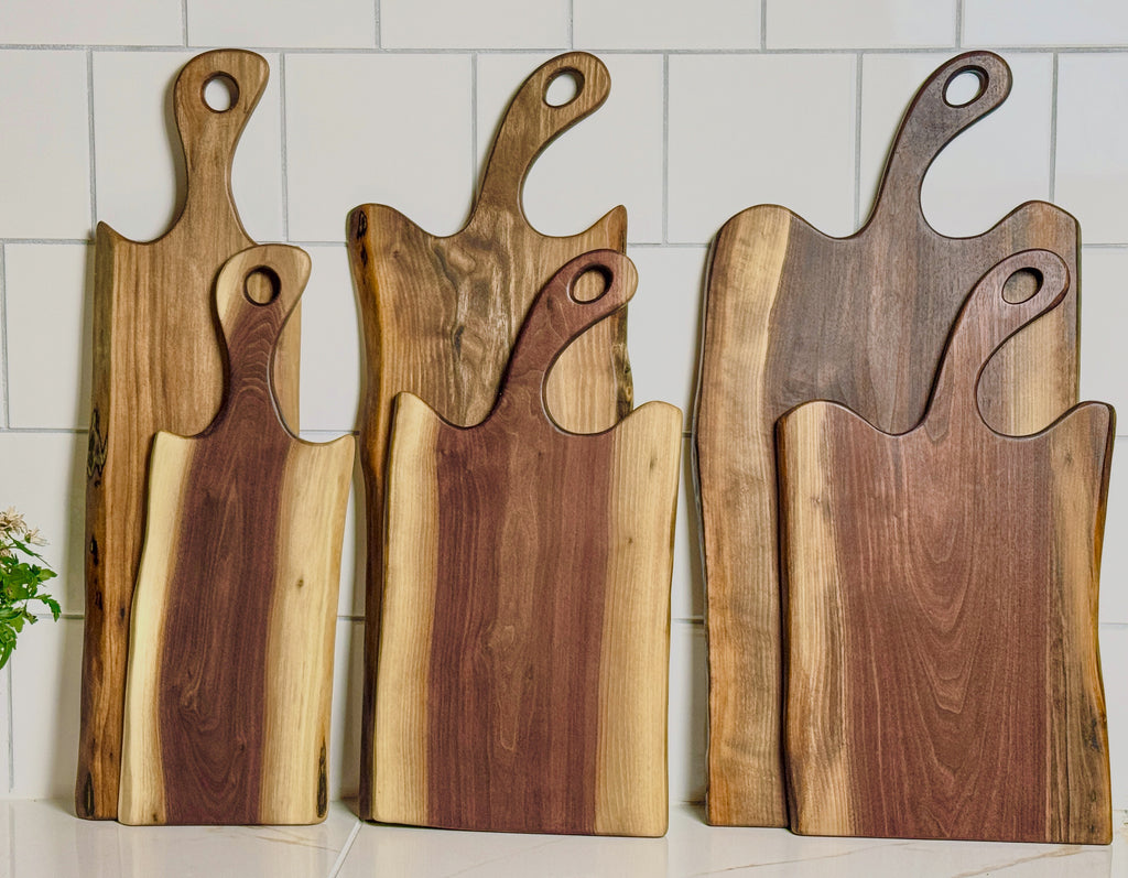 A selection of 6 live edge Black walnut charcuterie boards in varying heights and widths. Two of the boards are Sapwood, four boards are heartwood. The image showcases the variance from board to board.