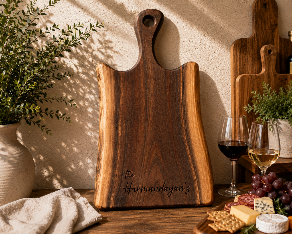 A dark wood cutting board with a handle and personalized with 'The Harmanfeyan's' stands on a wooden table in a styled kitchen or bar setting. Wine glasses, grapes, cheese, crackers, and other wooden boards are arranged around it against a textured beige wall with sunlight and leaf shadows.